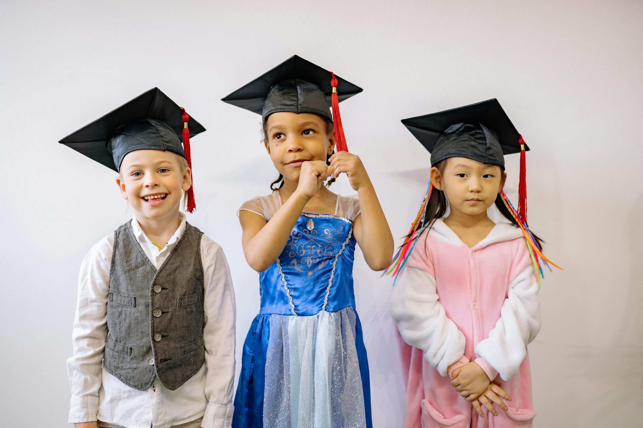 Three children in graduation caps express pride and joy at their preschool graduation ceremony.
